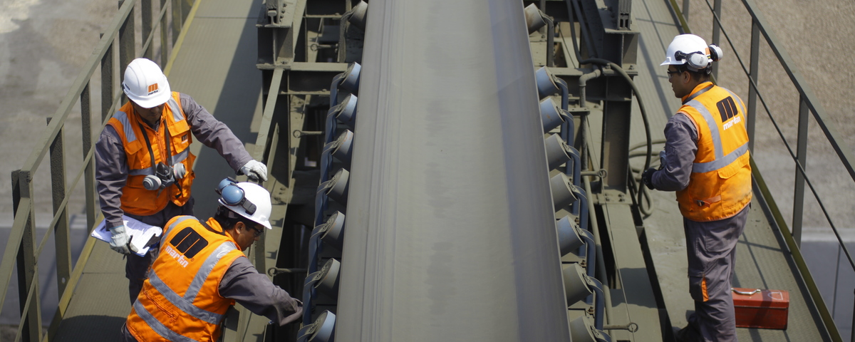 Երեք աշխատող նարնջագույն անվտանգության բաճկոններով և սպիտակ սաղավարտներով՝ կատարում արդյունաբերական փոխադրիչ ժապավենի համակարգի պահպանում: