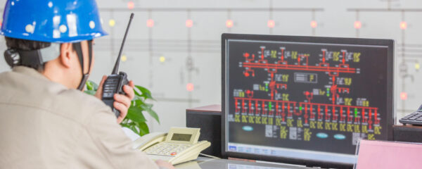 Արդյունաբերական աշխատող կապույտ պաշտպանիչ կասկետով՝ օգտագործում ռադիոկապ և վերահսկում կառավարման համակարգի էկրանը՝ ցուցադրելով օբյեկտի սխեմաները և կարգավիճակի ցուցիչները: