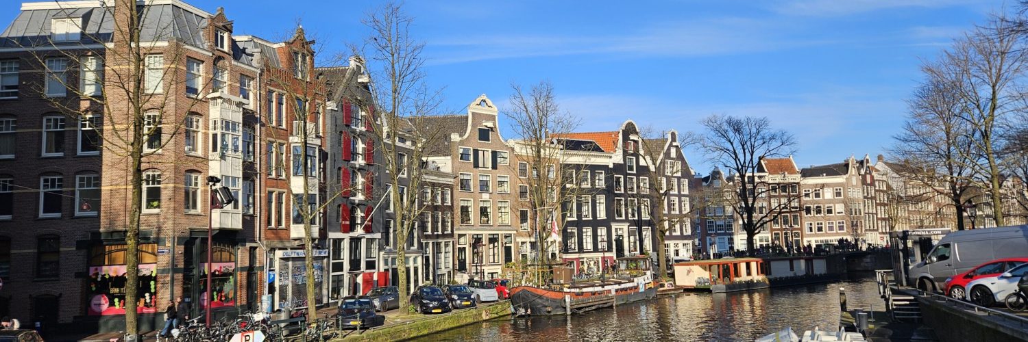 Armenian:
Ամստերդամի ջրանցքային տեսարան ավանդական հոլանդական շարքային տներով, ջրուղու երկայնքով կանգնած նավակներով, փողոցում կայանված հեծանիվներով և պայծառ կապույտ երկնքի տակ ցերեկային ծառերով